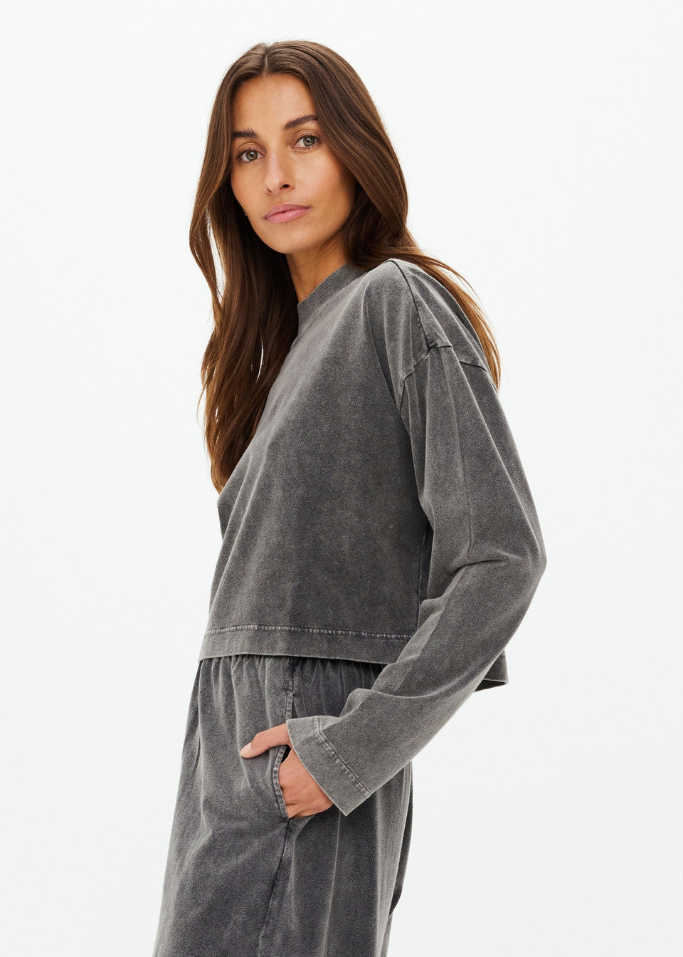 A woman with long brown hair wears a faded gray SOHO CROPPED LONG SLEEVE TOP and recycled cotton pants, one hand in her pocket, gazing neutrally at the camera against a plain white background. Brand: The Upside.