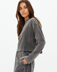 A woman with long brown hair wears a faded gray SOHO CROPPED LONG SLEEVE TOP and recycled cotton pants, one hand in her pocket, gazing neutrally at the camera against a plain white background. Brand: The Upside.