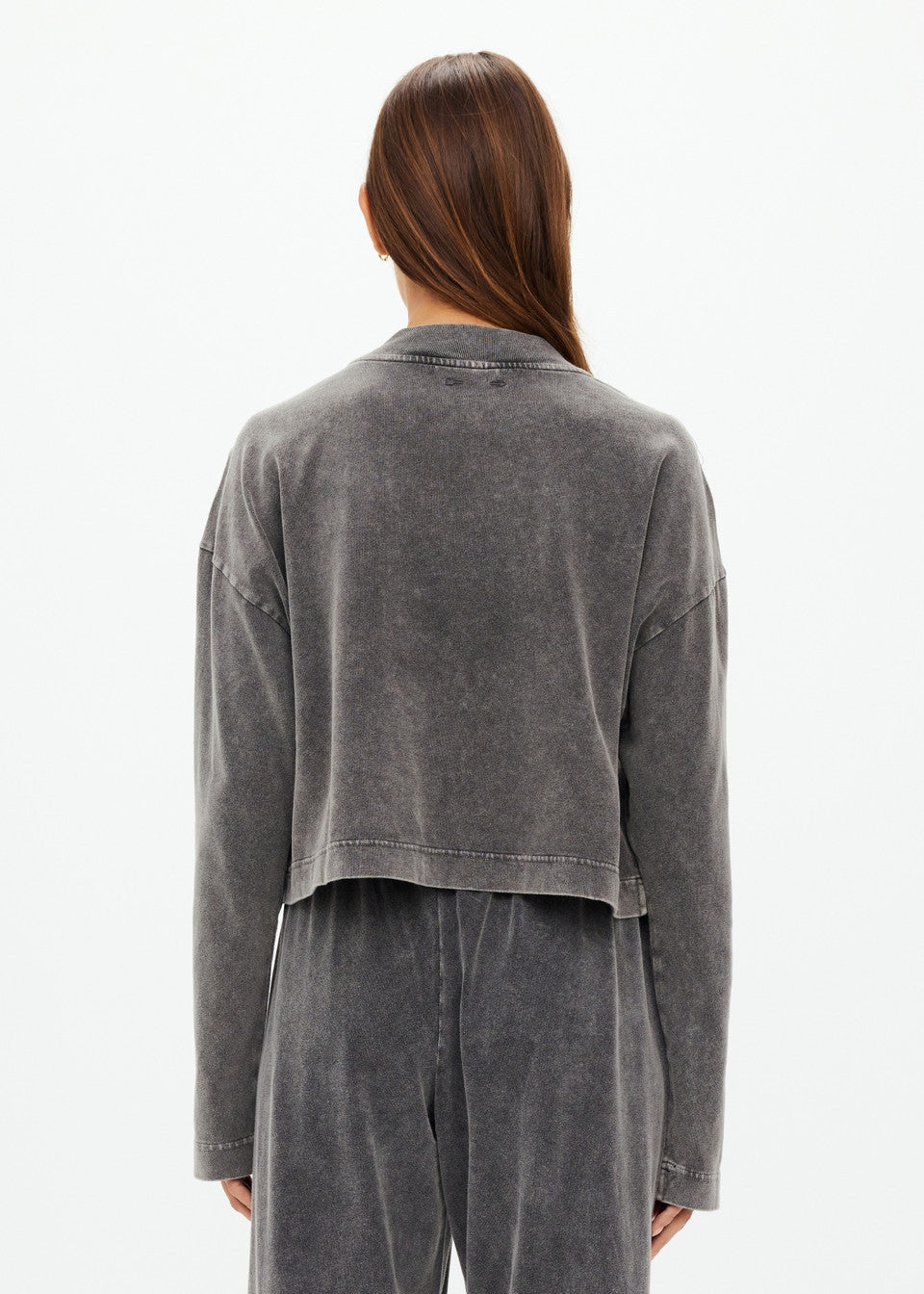 A person with long brown hair is wearing The Upside's SOHO CROPPED LONG SLEEVE TOP in faded dark gray, alongside matching pants, both made from recycled cotton. The background is plain light gray.