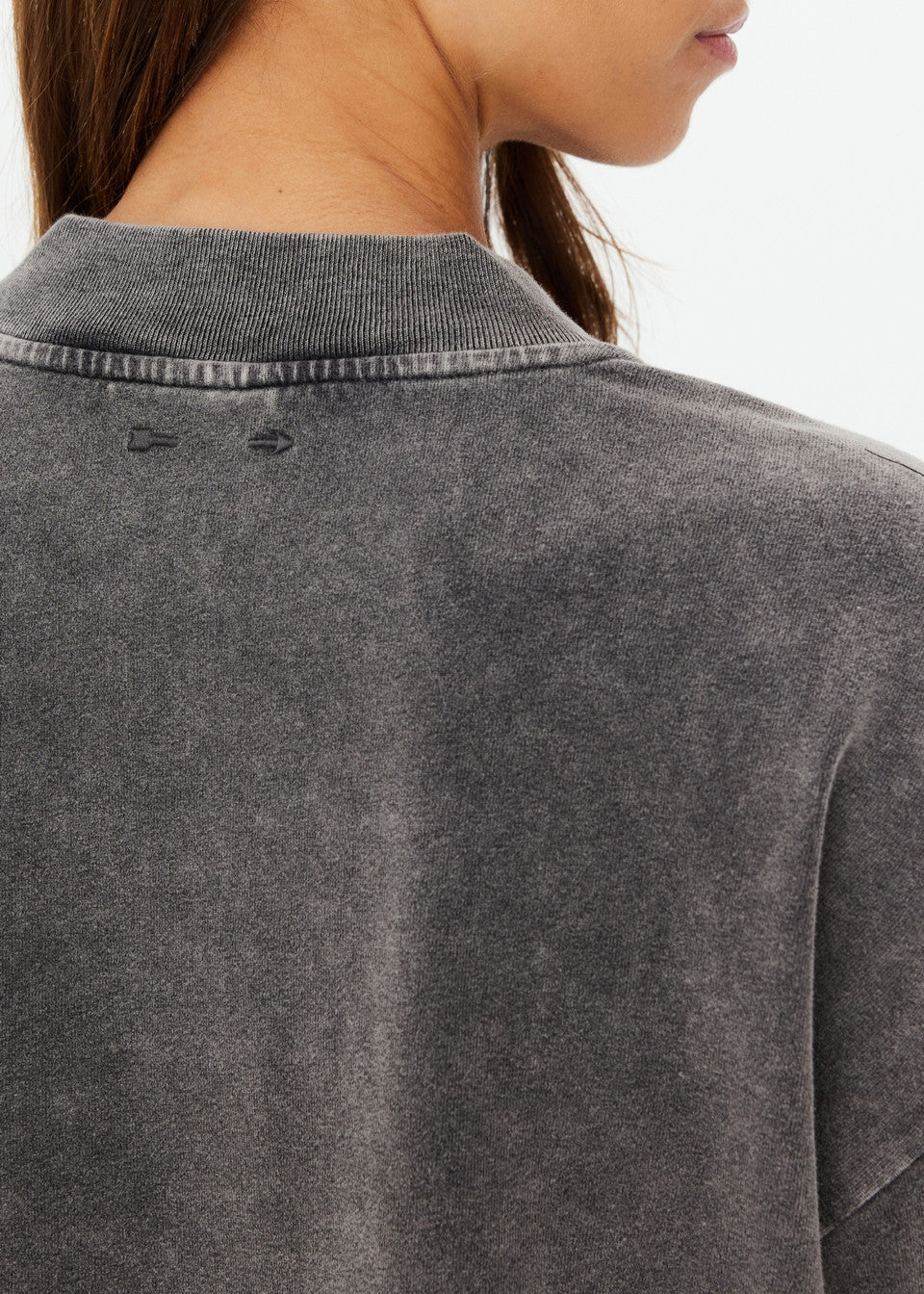 Close-up of someone wearing The Upside's SOHO Cropped Long Sleeve Top, made from recycled cotton with a ribbed round collar. The faded black shirt features a subtle distressed texture, partially concealed by their long brown hair from the back view.