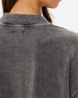 Close-up of someone wearing The Upside's SOHO Cropped Long Sleeve Top, made from recycled cotton with a ribbed round collar. The faded black shirt features a subtle distressed texture, partially concealed by their long brown hair from the back view.