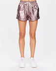 Against a plain white backdrop, a person wears The Upside's BILLIE RUN SHORTS in metallic pink with an elasticated waistband and white drawstring. The shiny, crinkled texture of the mid-rise shorts pairs perfectly with their crisp white sneakers.