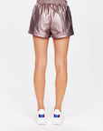 A person is seen from behind wearing The Upside's BILLIE RUN SHORTS in a metallic gunmetal finish with an elasticated waistband, paired with white sneakers featuring blue and red accents, standing against a plain white background.