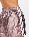 A close-up shows someone in The Upside's BILLIE RUN SHORTS, metallic pink with a crinkled texture. These mid-rise, lightweight shorts have an elastic waistband, white-striped sides, and a gunmetal finish against a plain white background.