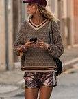 A person in a patterned sweater, The Upside's BILLIE RUN SHORTS with an elasticated waistband, and a maroon cap walks down the street holding a phone. They carry a crossbody bag and look to the side against blurred buildings.