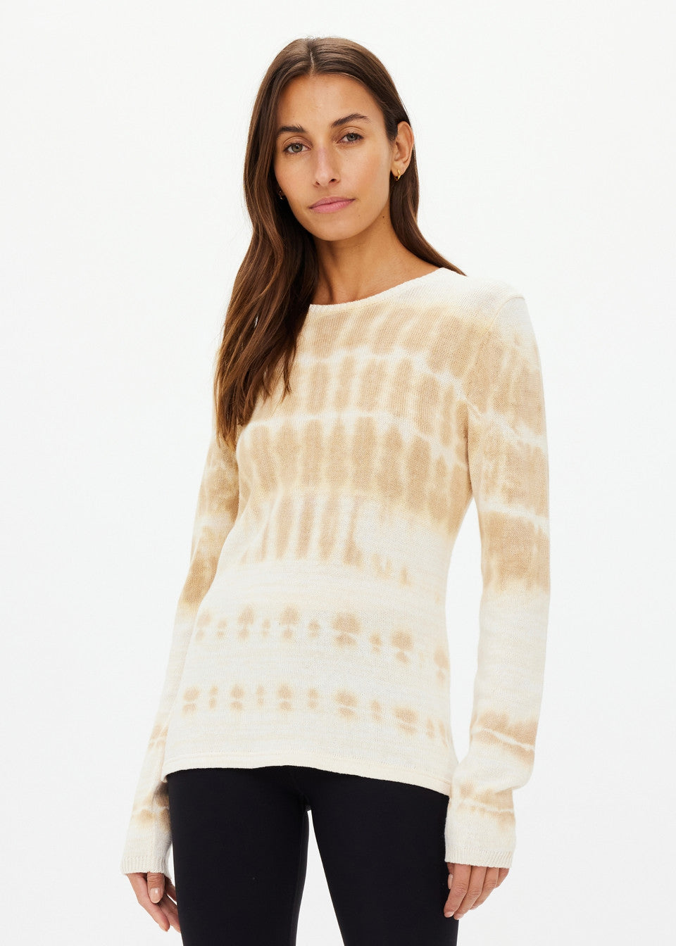 A person with long brown hair is wearing The Upside's AMANA LONG SLEEVE KNIT TOP, hand tie-dyed and made from organic cotton, paired with black leggings, standing against a plain white background.