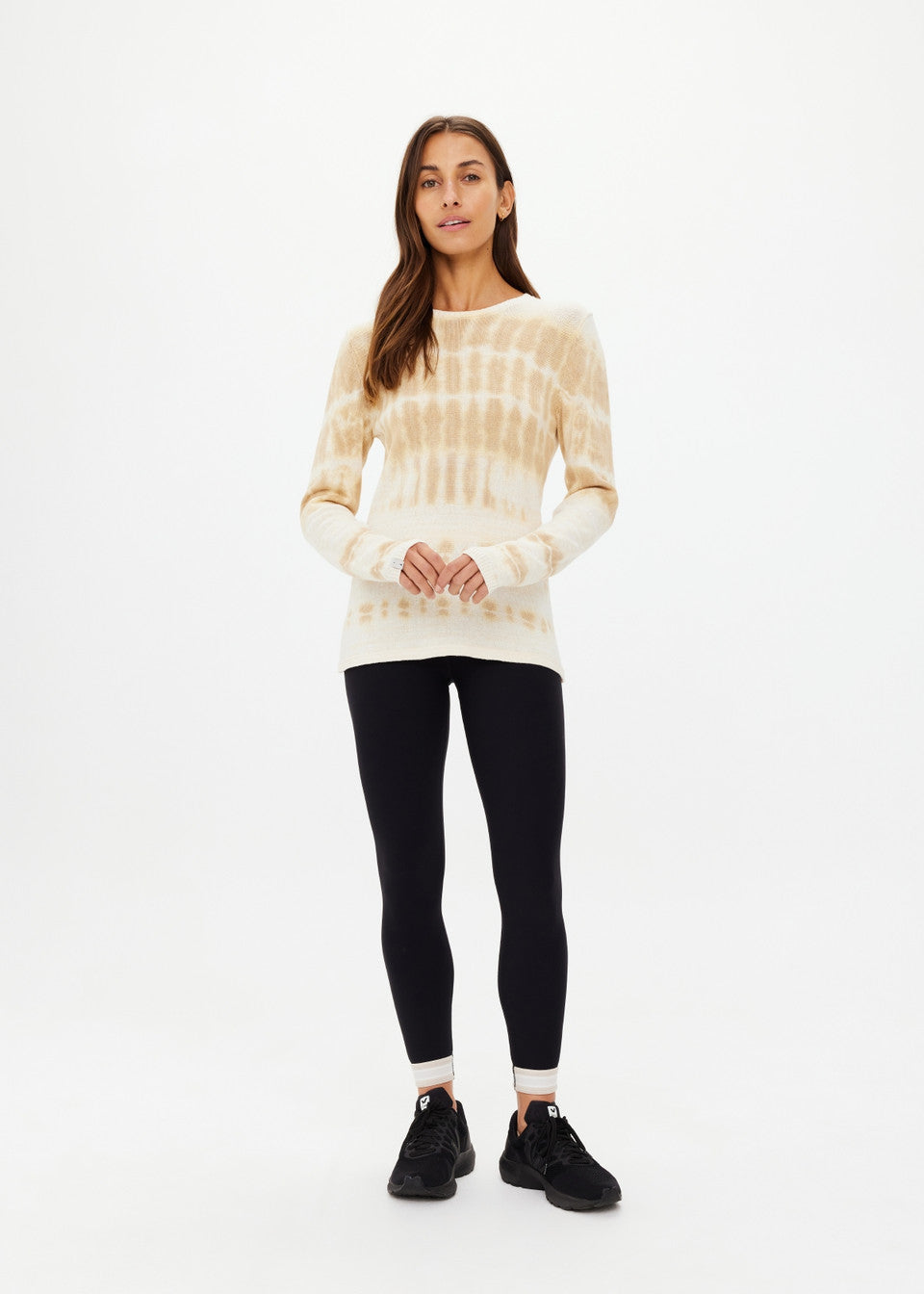 A woman with long hair stands against a plain background, wearing The Upside's beige tie-dye AMANA LONG SLEEVE KNIT TOP made of organic cotton and black leggings. Her top pairs effortlessly with her black sneakers as she loosely clasps her hands in front.