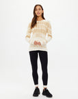 A woman with long hair stands against a plain background, wearing The Upside's beige tie-dye AMANA LONG SLEEVE KNIT TOP made of organic cotton and black leggings. Her top pairs effortlessly with her black sneakers as she loosely clasps her hands in front.