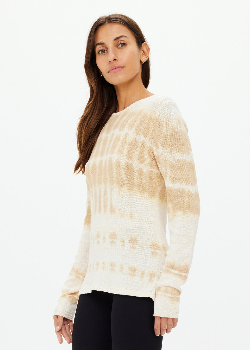 A woman poses against a plain background, showcasing The Upside's AMANA LONG SLEEVE KNIT TOP in beige and white tie-dye. Her long brown hair flows elegantly as she looks to the side, pairing the organic cotton top with chic black pants.