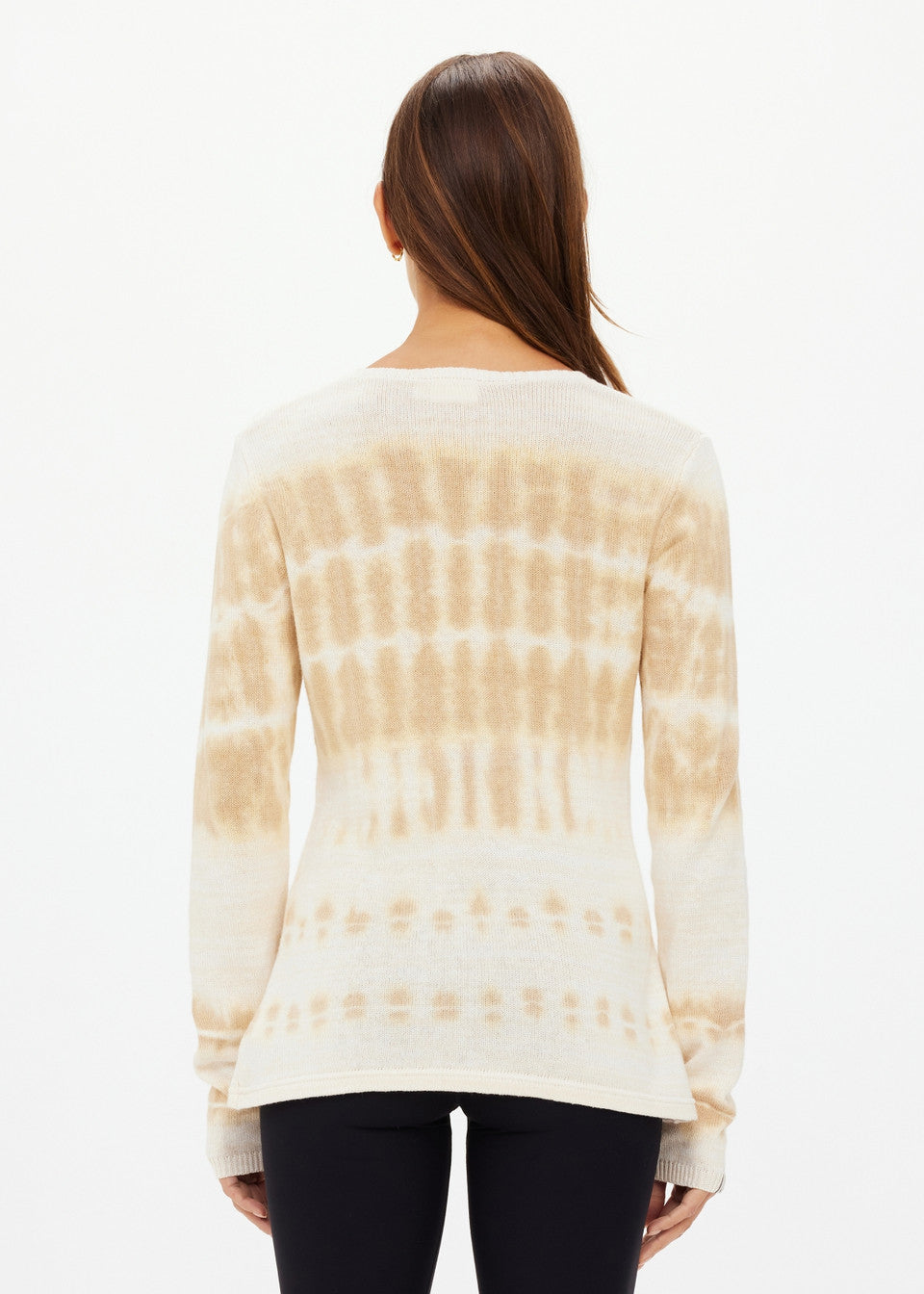 A person with long brown hair stands against a white background, wearing an AMANA LONG SLEEVE KNIT TOP by The Upside, featuring a vertical tie-dye pattern in varying shades of beige, paired with black leggings.