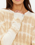 A person in The Upside's AMANA LONG SLEEVE KNIT TOP in a beige and white hand-dyed design gently holds the collar. The sweater's soft texture complements their long brown hair, with only the lower part of their face visible.