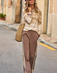 A woman strolls down the street in The Upside's AMANA LONG SLEEVE KNIT TOP, flaunting vibrant hand tie-dyed colors. Her brown trousers with white stripes and woven bag enhance her look, while a purple headscarf and sunglasses add flair against a building with a stone facade.