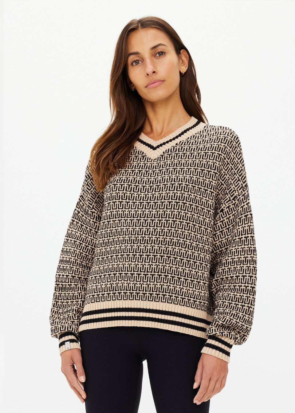 A person with long brown hair is wearing The Upside's CASTILLA MILLIE KNIT SWEATER, a patterned knitted sweater made from organic cotton with black and beige geometric designs, ribbed cuffs, and a V-neckline. They're paired with black pants against a plain white background.