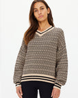 A person with long brown hair is wearing The Upside's CASTILLA MILLIE KNIT SWEATER, a patterned knitted sweater made from organic cotton with black and beige geometric designs, ribbed cuffs, and a V-neckline. They're paired with black pants against a plain white background.