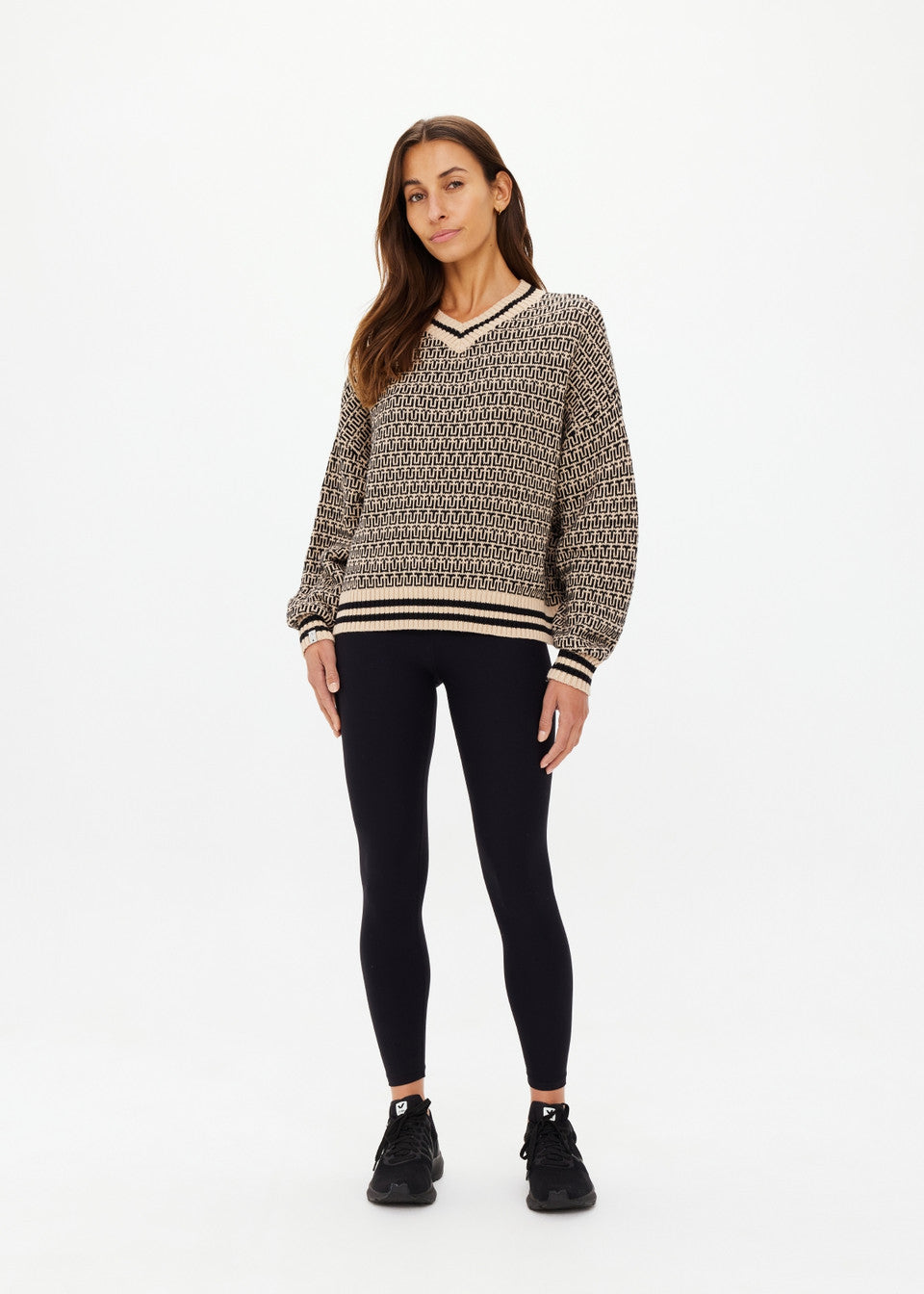 A woman with long brown hair wears The Upside CASTILLA MILLIE KNIT SWEATER, along with black leggings and sneakers. She stands against a plain white background, looking directly at the camera with a neutral expression.