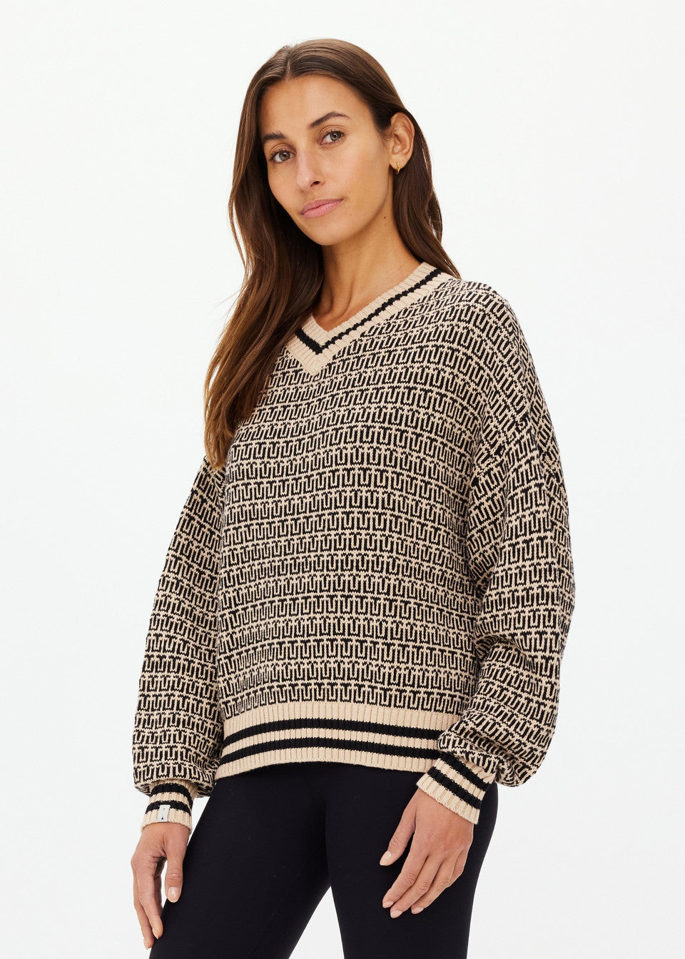 A woman with long brown hair stands against a plain white background wearing The Upside's CASTILLA MILLIE KNIT SWEATER. It features long sleeves and a V-neck and is crafted from organic cotton with a geometric beige and black design. She looks towards the camera.