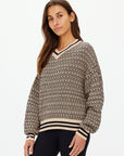 A woman with long brown hair stands against a plain white background wearing The Upside's CASTILLA MILLIE KNIT SWEATER. It features long sleeves and a V-neck and is crafted from organic cotton with a geometric beige and black design. She looks towards the camera.