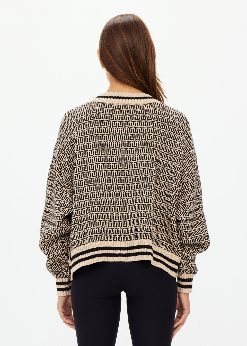 Rear view of a person with long hair wearing The Upside CASTILLA MILLIE KNIT SWEATER, crafted from organic cotton, showcasing a black and beige geometric design. They pair it with black pants against a plain white background.