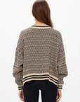 Rear view of a person with long hair wearing The Upside CASTILLA MILLIE KNIT SWEATER, crafted from organic cotton, showcasing a black and beige geometric design. They pair it with black pants against a plain white background.