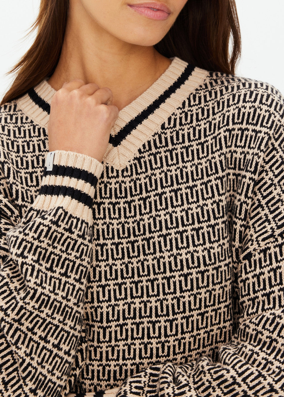 A person wears The Upside's CASTILLA MILLIE KNIT SWEATER in beige, featuring a black geometric pattern and striped trim at the neckline and cuffs. Made from organic cotton, their hand rests near the collar against a simple background.