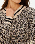 A person wears The Upside's CASTILLA MILLIE KNIT SWEATER in beige, featuring a black geometric pattern and striped trim at the neckline and cuffs. Made from organic cotton, their hand rests near the collar against a simple background.