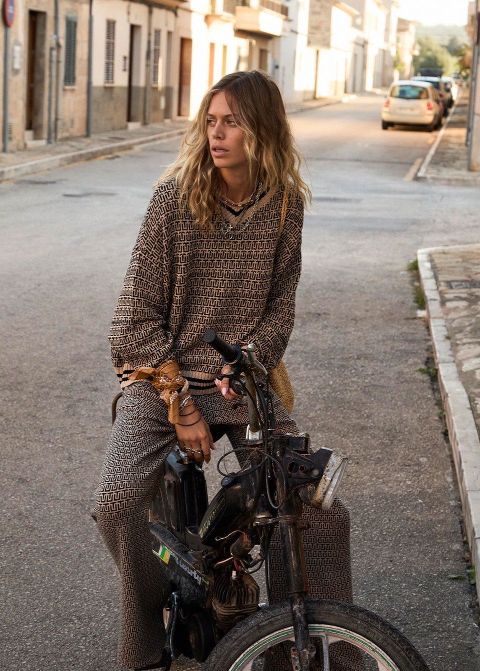 A person with long hair sits on a bicycle in a quiet street lined with buildings. They wear The Upside's CASTILLA MILLIE KNIT SWEATER, featuring a subtle monogram design, as they gaze to the side. Parked cars are visible in the background.