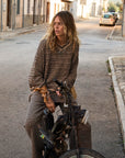 A person with long hair sits on a bicycle in a quiet street lined with buildings. They wear The Upside's CASTILLA MILLIE KNIT SWEATER, featuring a subtle monogram design, as they gaze to the side. Parked cars are visible in the background.