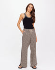 A woman with long brown hair poses against a plain white background, wearing a black sleeveless top and The Upside's Castilla Juniper Knit Pant. Her gray patterned wide-leg pants showcase a sleek knit jacquard design made from an organic cotton blend. She also wears black sandals and has one hand in her pocket.