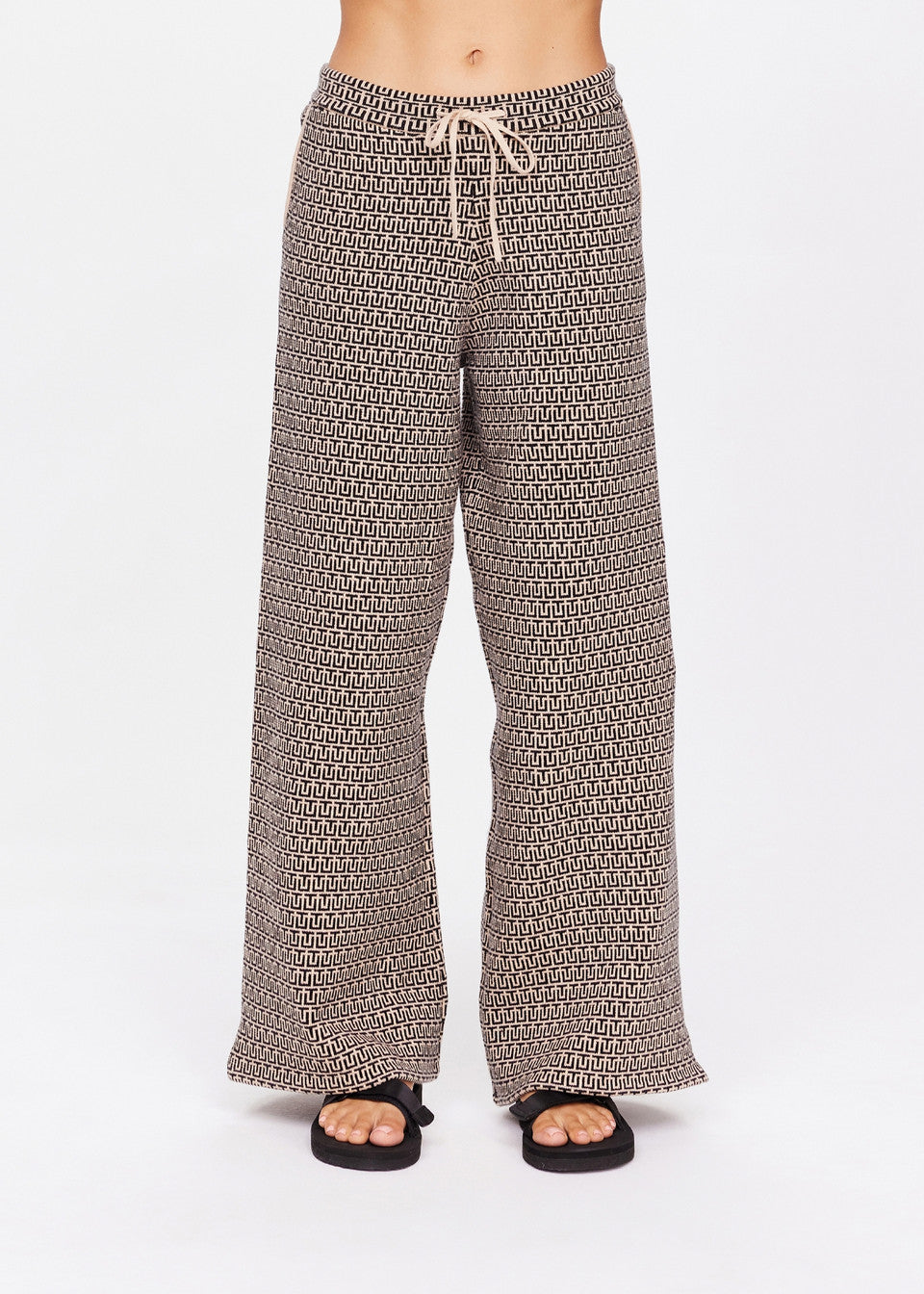 A person is wearing The Upside's Castilla Juniper Knit Pant, featuring a small repeating pattern and drawstring waistband, paired with black sandals. The photo highlights the loose fit and flowy organic cotton blend fabric against a plain background, focusing on the lower half of the body.