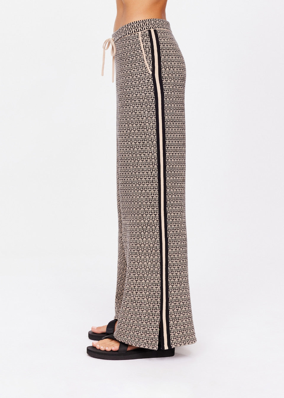 A person is seen from the side wearing The Upside's Castilla Juniper Knit Pant featuring a geometric pattern, drawstring waist, and wide-leg design. It has a vertical black stripe along the side. They complete the look with black sandals against a plain white backdrop.