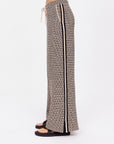 A person is seen from the side wearing The Upside's Castilla Juniper Knit Pant featuring a geometric pattern, drawstring waist, and wide-leg design. It has a vertical black stripe along the side. They complete the look with black sandals against a plain white backdrop.