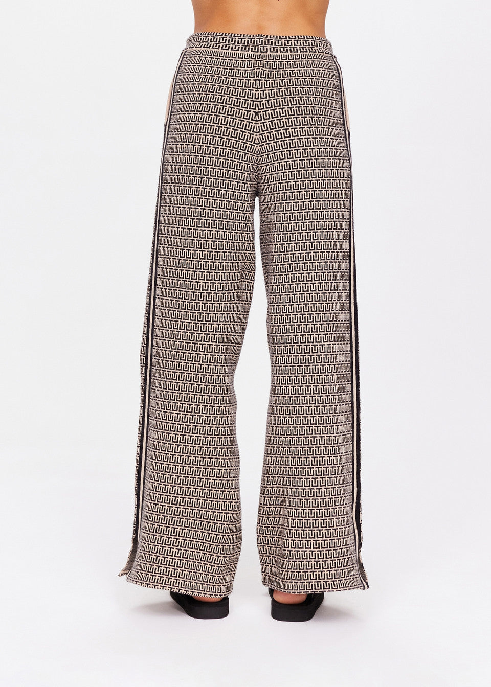 A person is wearing The Upside's Castilla Juniper Knit Pant featuring a black and cream geometric design. This jacquard piece includes side pockets and bold black vertical leg stripes against a white background with black shoes.