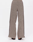 A person is wearing The Upside's Castilla Juniper Knit Pant featuring a black and cream geometric design. This jacquard piece includes side pockets and bold black vertical leg stripes against a white background with black shoes.