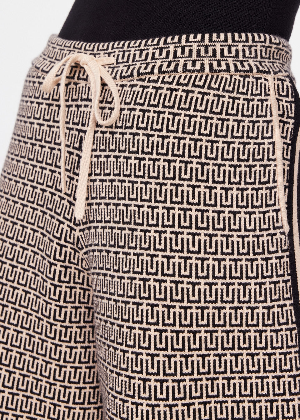 A close-up showcases The Upside's CASTILLA JUNIPER KNIT PANT, featuring a geometric jacquard pattern in black and beige. Made from an organic cotton blend, these pants include a drawstring waistband.