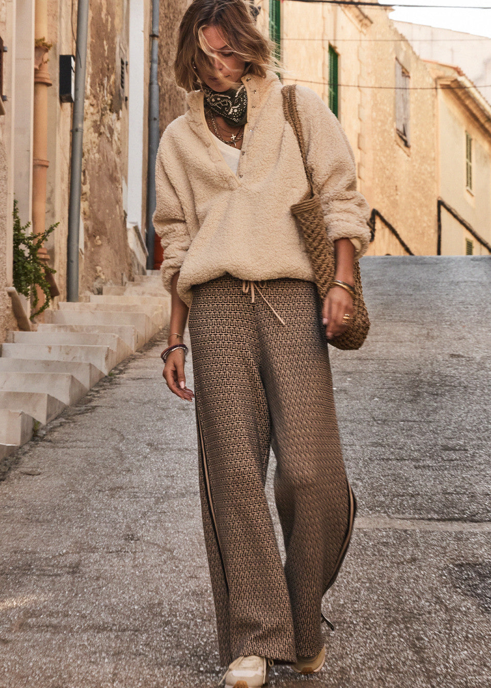 Wandering a quaint street, a person wears The Upside's CASTILLA JUNIPER KNIT PANT and a beige fleece pullover, carrying a woven bag with bracelets. A scarf graces their neck as stone steps and historic facades create an enchanting backdrop to their stroll.