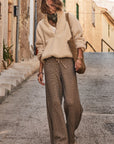 Wandering a quaint street, a person wears The Upside's CASTILLA JUNIPER KNIT PANT and a beige fleece pullover, carrying a woven bag with bracelets. A scarf graces their neck as stone steps and historic facades create an enchanting backdrop to their stroll.