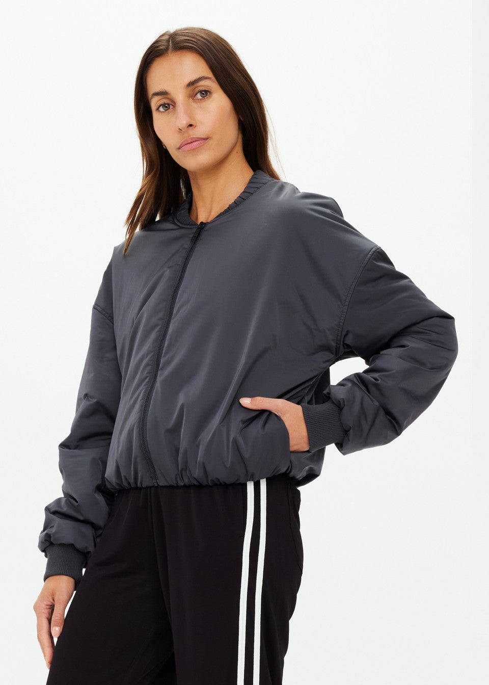 A person with long brown hair wears The Upside's CARLOTTA CROPPED BOMBER JACKET made of recycled polyester, featuring an embossed apple leather patch, and black pants with white stripes. They stand against a plain white background, looking directly at the camera with a neutral expression.