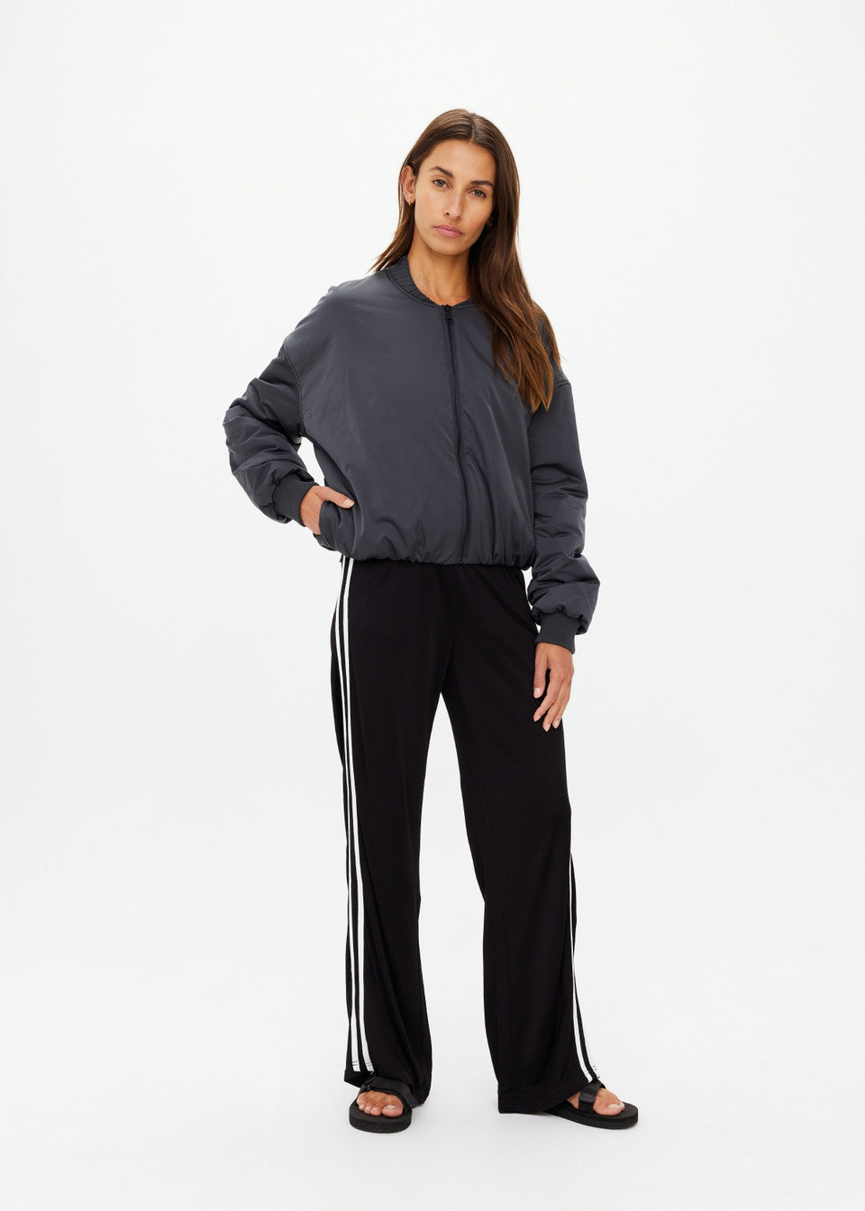 A woman with long hair confidently poses in The Upside's CARLOTTA CROPPED BOMBER JACKET featuring an embossed apple leather patch, black track pants with white stripes, and black sandals. She has one hand in her pocket against a plain white background.