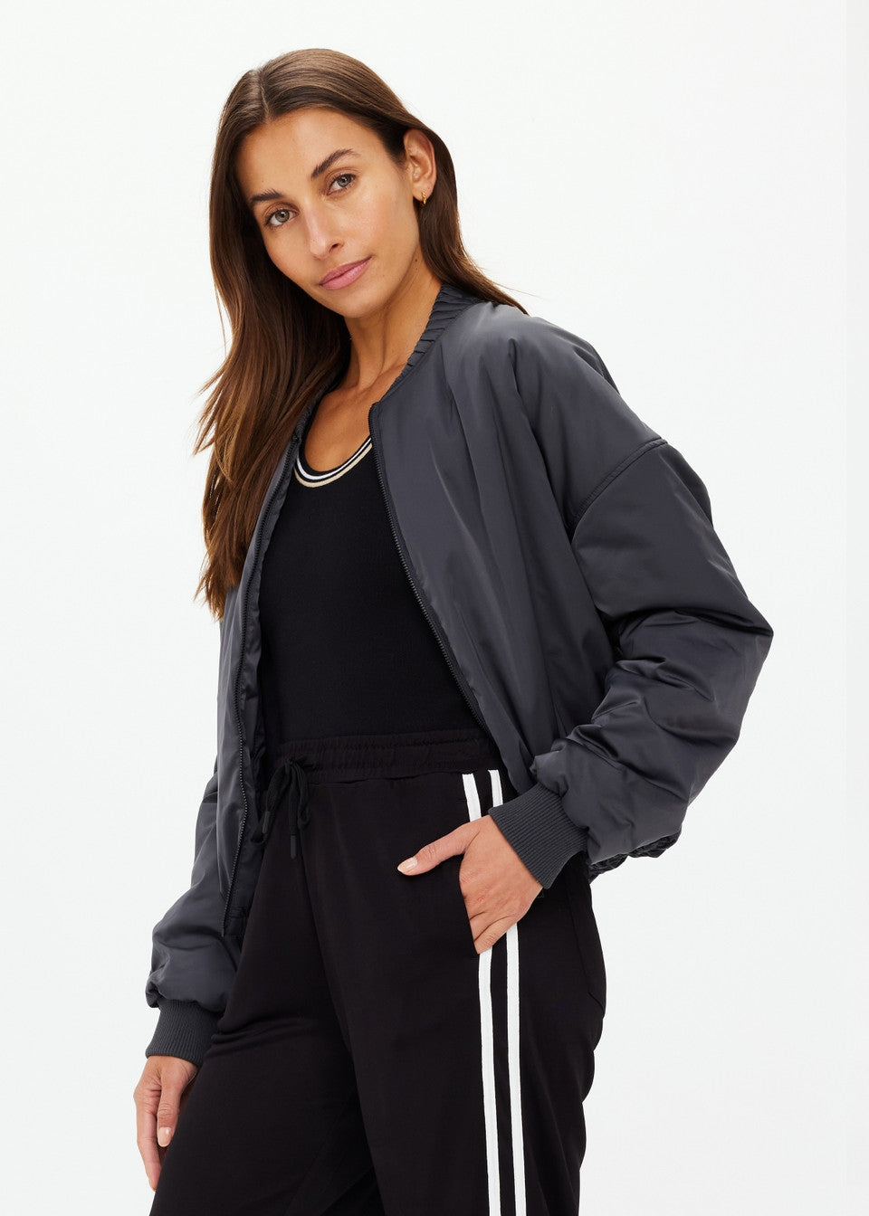 A person with long, dark hair wears The Upside's CARLOTTA CROPPED BOMBER JACKET in black, made from recycled polyester, paired with black sweatpants featuring white stripes. They stand against a plain white background, looking at the camera neutrally.