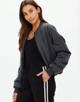 A person with long, dark hair wears The Upside's CARLOTTA CROPPED BOMBER JACKET in black, made from recycled polyester, paired with black sweatpants featuring white stripes. They stand against a plain white background, looking at the camera neutrally.