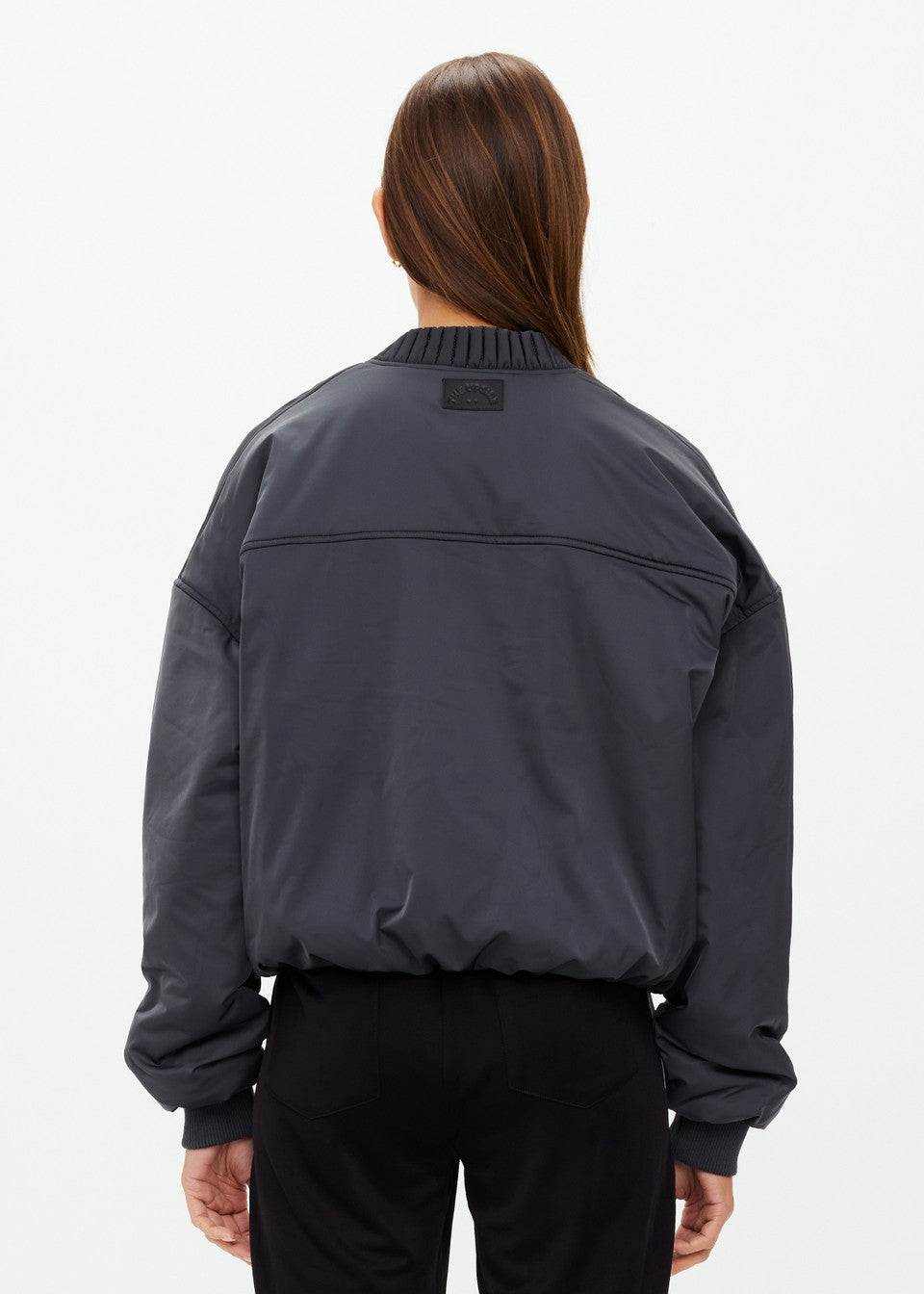 A person with long brown hair wears The Upside's dark gray CARLOTTA CROPPED BOMBER JACKET with an embossed apple leather patch, facing away from the camera against a neutral background. Its high collar and ribbed cuffs contribute to its unique style.