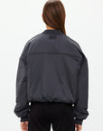 A person with long brown hair wears The Upside's dark gray CARLOTTA CROPPED BOMBER JACKET with an embossed apple leather patch, facing away from the camera against a neutral background. Its high collar and ribbed cuffs contribute to its unique style.