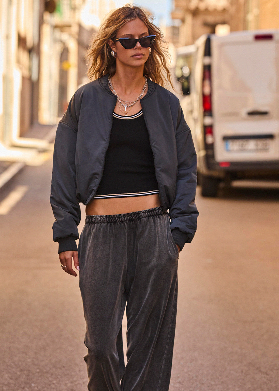 A person wearing sunglasses and The Upside's Carlotta Cropped Bomber Jacket, crafted from recycled polyester with an adjustable fit, confidently strolls down a sunny street in gray pants, while a white van is parked in the background.