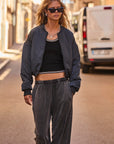 A person wearing sunglasses and The Upside's Carlotta Cropped Bomber Jacket, crafted from recycled polyester with an adjustable fit, confidently strolls down a sunny street in gray pants, while a white van is parked in the background.