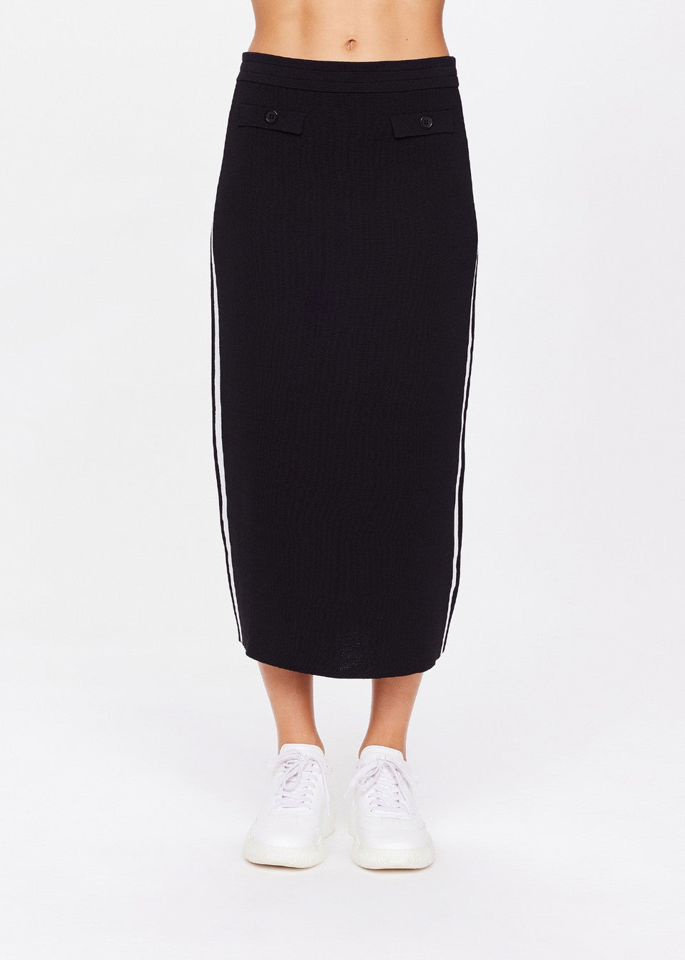 A person models The Upside's ATOMIC KNIT MIDI SKIRT, a long black piece with white side stripes and front buttons, paired with white sneakers. A plain white background accentuates the elegant texture contrast from waist to feet.