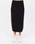 A person models The Upside's ATOMIC KNIT MIDI SKIRT, a long black piece with white side stripes and front buttons, paired with white sneakers. A plain white background accentuates the elegant texture contrast from waist to feet.