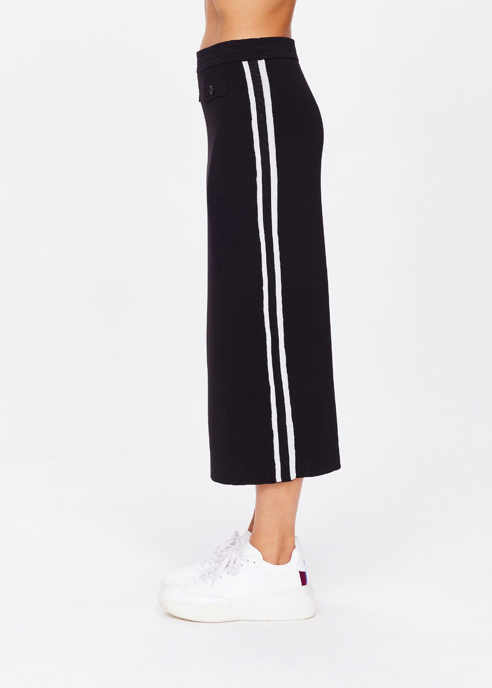 Wearing The Upside's ATOMIC KNIT MIDI SKIRT, featuring white vertical stripes on a long black design with an elastic waistband, paired with white sneakers. The side profile against a plain white background exudes effortless style.