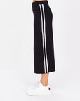 Wearing The Upside's ATOMIC KNIT MIDI SKIRT, featuring white vertical stripes on a long black design with an elastic waistband, paired with white sneakers. The side profile against a plain white background exudes effortless style.