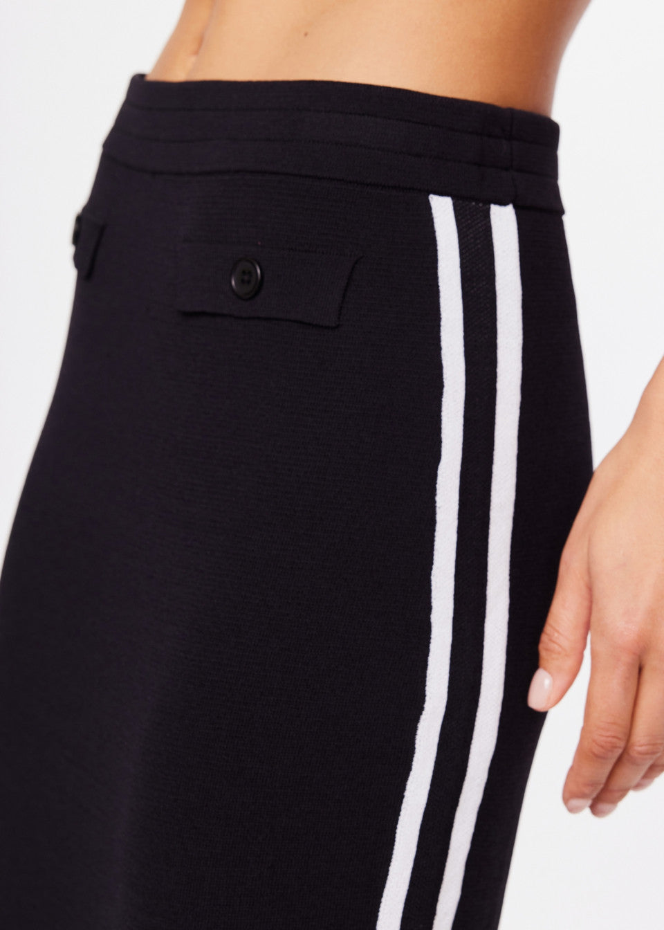 Close-up of someone wearing The Upside's black ATOMIC KNIT MIDI SKIRT, featuring two white side stripes and buttons near the elastic waistband; hand visible.