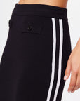 Close-up of someone wearing The Upside's black ATOMIC KNIT MIDI SKIRT, featuring two white side stripes and buttons near the elastic waistband; hand visible.